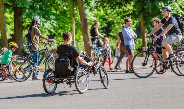 A photo of many people walking, riding their bikes or using a wheel chair. They are on a path surrounded by trees.